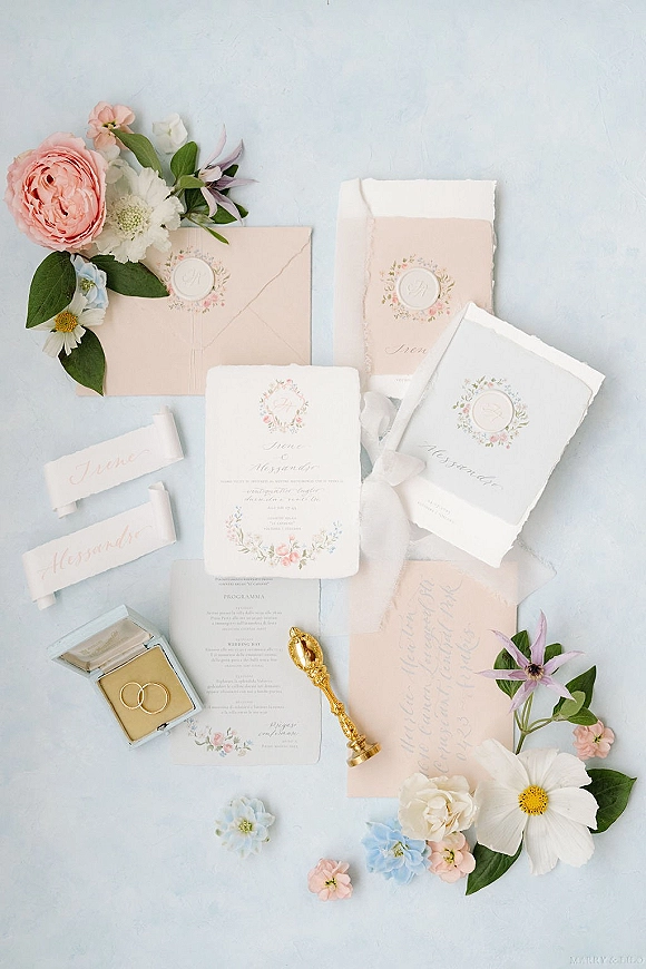 Invitation suite wedding invitation flat lay with blush pink envelope, deckle edge cards, floral wreath, wax seal, and rings on light blue backdrop