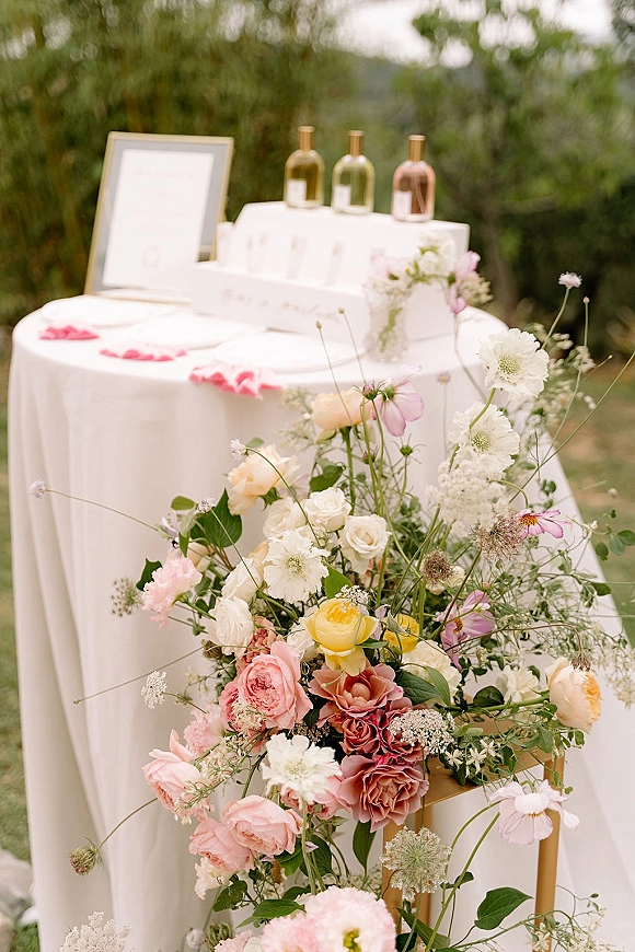 Wedding welcome table with wedding welcome sign in a frame, garden rose florals, bud vases, perfume bottles, and rose petals outdoors