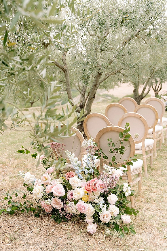 Ceremony seating with outdoor wedding chairs lined by pink and white garden rose aisle florals on a lawn beneath olive trees and hills