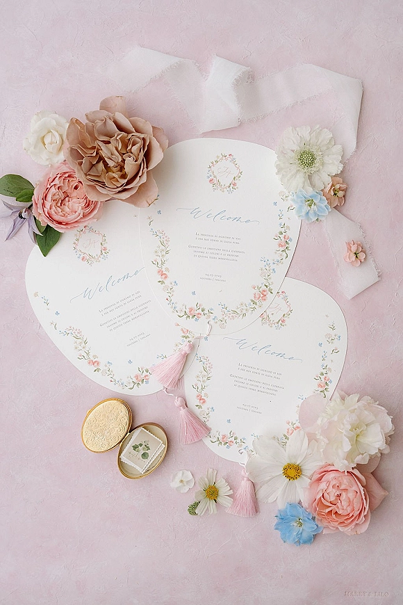 Wedding invitations with floral wreath invitation design, calligraphy text, pink tassels, wax-sealed envelopes, flowers, and gold oval ring box on a pink textured surface