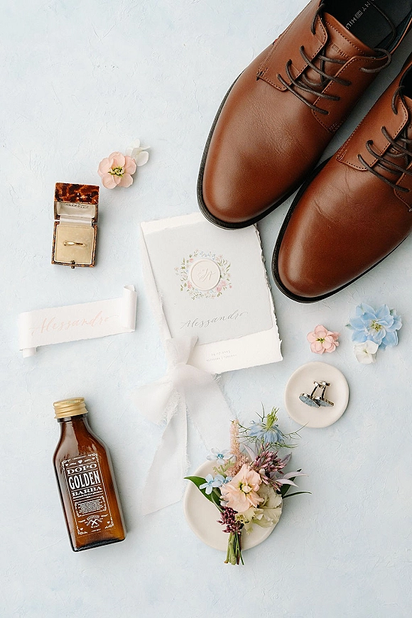 Wedding flat lay of brown leather dress shoes, wedding invitation with wax seal, rings and boutonniere on a light blue textured surface