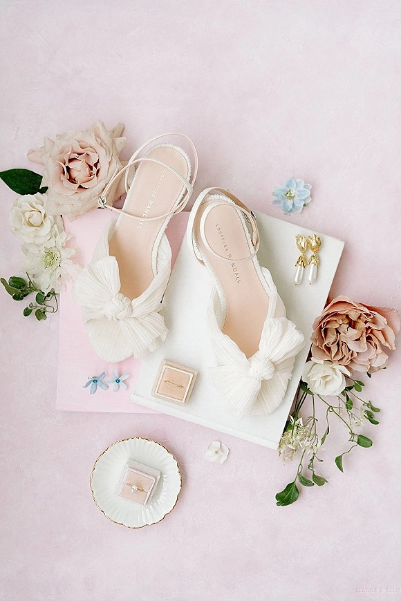 Bridal details flatlay with wedding details flat lay of white bow shoes, earrings, engagement ring, stationery, and florals on a pink backdrop