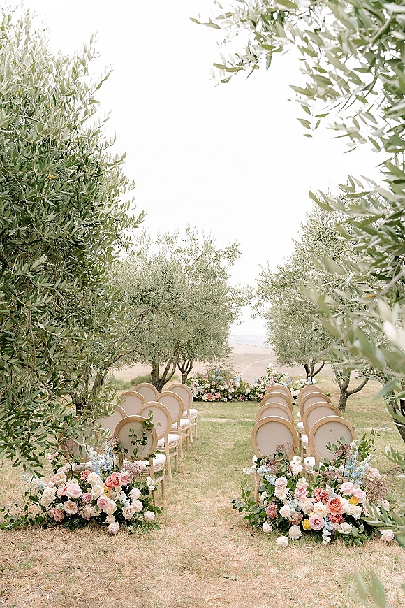 Ceremony aisle decor with outdoor ceremony aisle flowers lining upholstered oval-back chairs on a lawn beneath olive trees and open sky