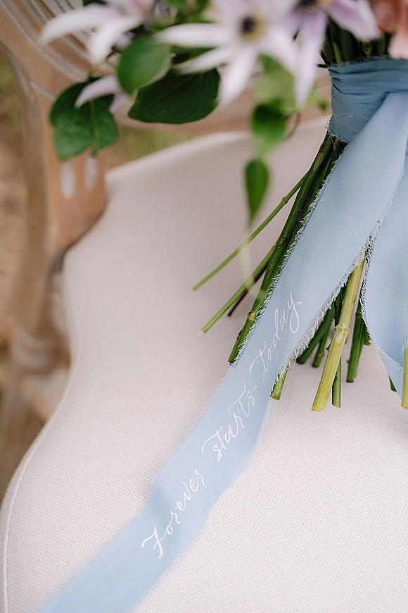 Bouquet detail with wedding bouquet ribbon in light blue, handwritten lettering trailing from white flowers and greenery on an upholstered chair