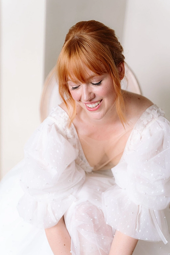 Bridal portrait of a redhead bride in a deep V wedding dress with sheer polka dot puff sleeves and veil against a white wall