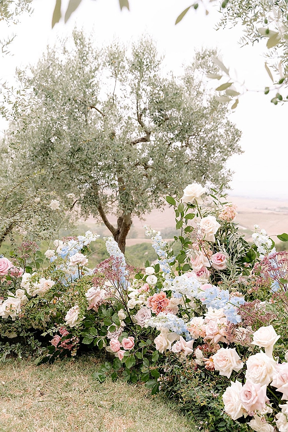 Wedding floral installation with ceremony ground florals, rose flowers lining a grassy aisle beneath an olive tree on a hillside lawn