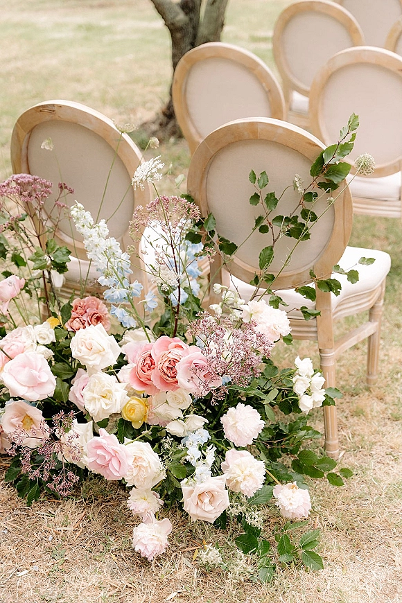 Ceremony aisle decor with aisle ground florals of pink and white roses and greenery beside upholstered wood-framed chairs on a lawn
