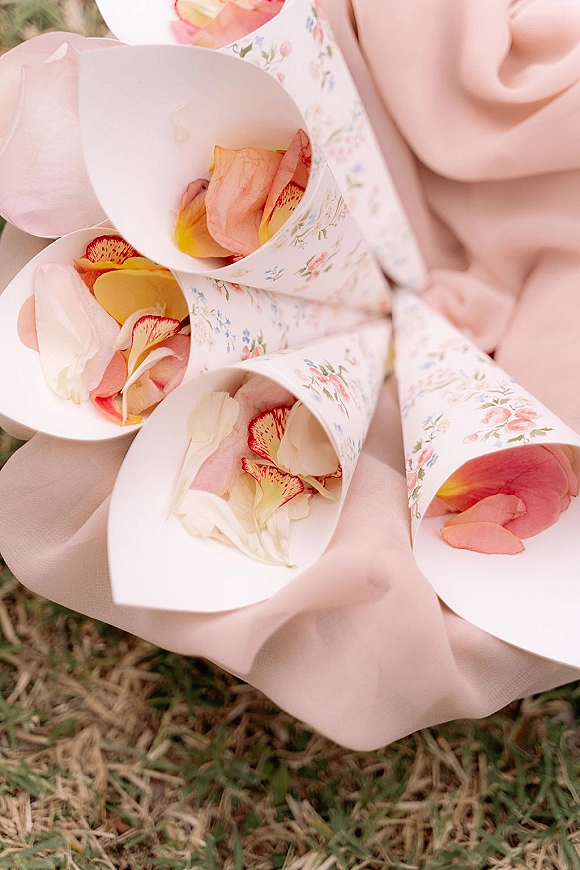 Flower petal cones and wedding confetti cones in floral print paper, filled with rose petals and set on blush fabric on grass