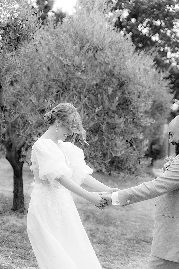First look moment outdoors as bride in puff sleeve wedding dress smiles holding groom’s hand on a garden lawn with trees behind