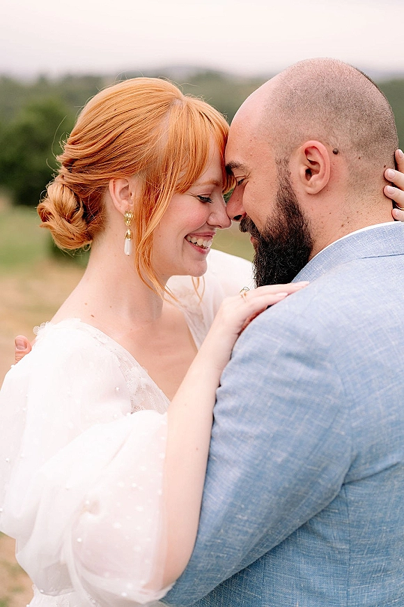 Couple portrait of bride in veil and wedding dress touching foreheads with bearded groom in light blue suit in an open field