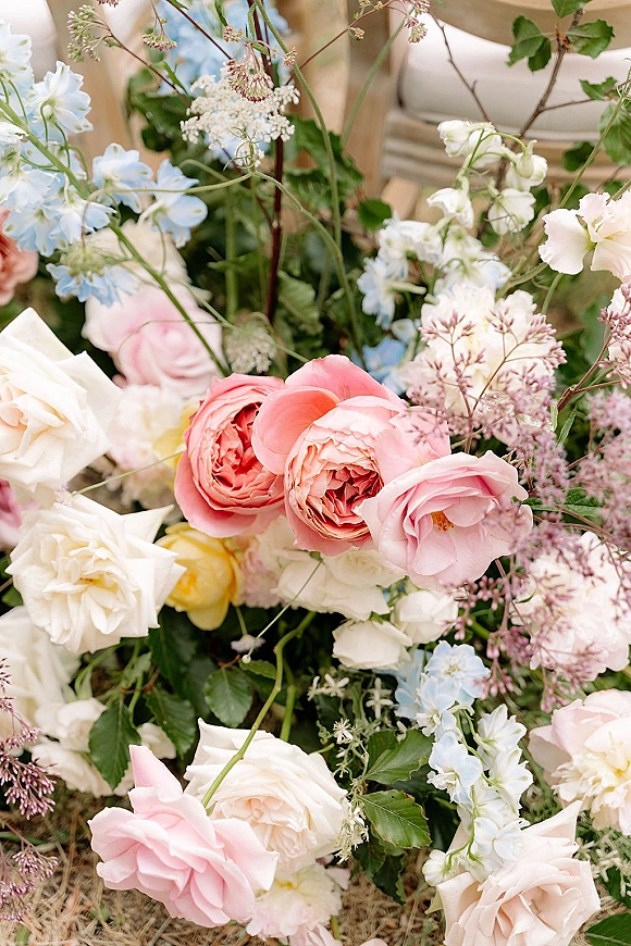 Wedding florals with a garden rose arrangement in blush, white, and yellow roses, blue delphinium, and greenery beside a wooden chair on grass lawn