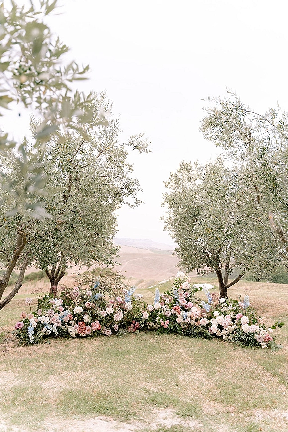 Ceremony floral arrangement of grounded ceremony flowers in pastel roses and delphinium lining an aisle, framed by olive trees on a grassy hill
