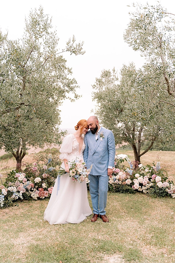Couple portrait of bride leaning on groom holding hands, her veil and bouquet visible, with olive trees and open sky behind