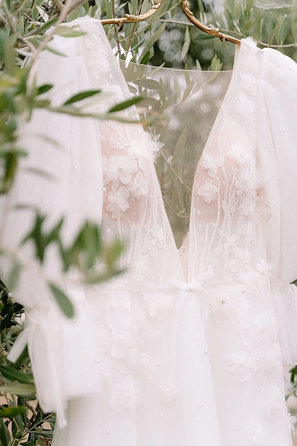 Wedding dress with lace applique and deep V neckline on a gold hanger, tulle overlay and pearl beading against green foliage backdrop