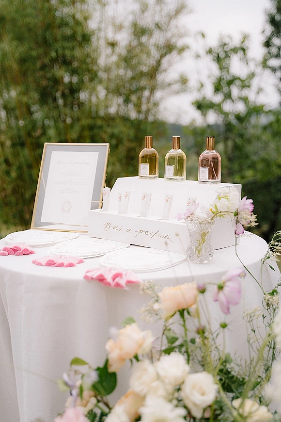 Wedding favor table with wedding perfume favors, framed sign, scent strips, favor boxes and floral arrangement on a round white table outdoors
