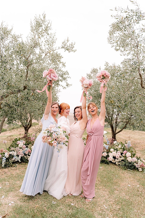 Bridesmaid group photo with bridesmaids holding bouquets, arms raised beside the bride on a lawn with olive trees and pink flowers