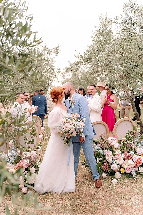 Wedding kiss as bride holds a pastel bouquet, groom in suit by ribboned aisle florals, with olive trees in the garden behind