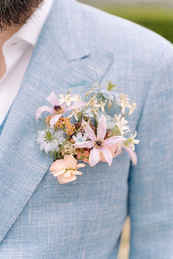 Wedding boutonniere on a light blue suit jacket with pink, white, and blue blooms and greenery against a softly blurred outdoor backdrop