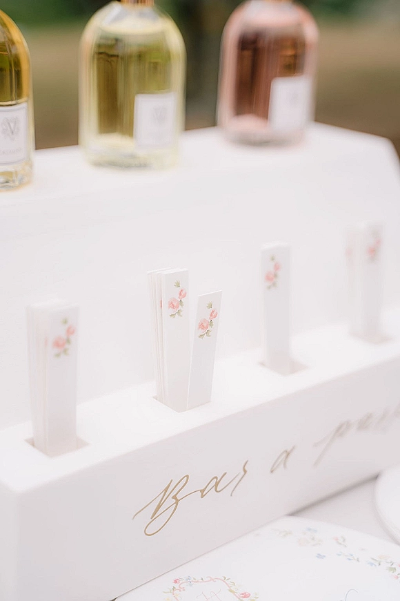 Wedding bar sign with signature drinks sign in calligraphy on clear acrylic, set on a white counter with wine bottles and greenery blur