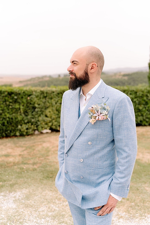 Groom portrait in a light blue groom suit, hands in pockets with pastel boutonniere and pocket square on a garden path backdrop