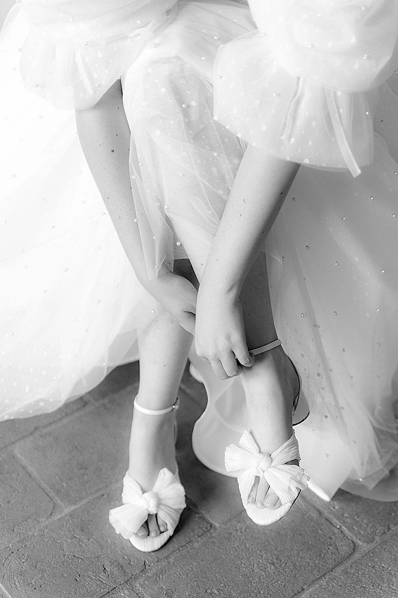 Bridal shoes with tulle wedding dress, white bow ankle-strap sandals and pearl beading on a stone floor beside a chair