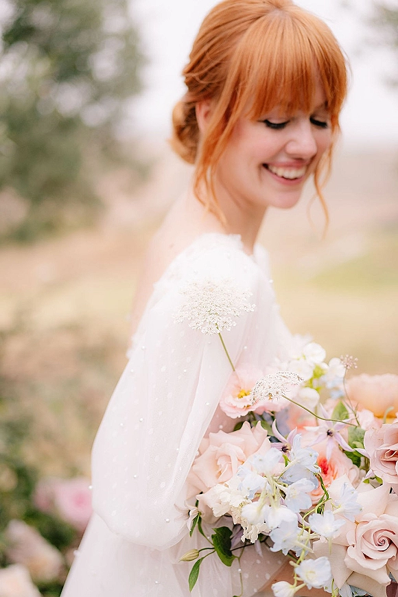 Bridal portrait of a smiling bride holding bouquet in an off-the-shoulder wedding dress and veil in an outdoor field with trees