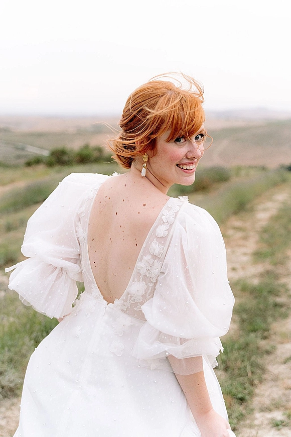 Bridal portrait of a redhead bride looking over her shoulder in a low-back lace gown with pearl accents in a grassy field under overcast sky
