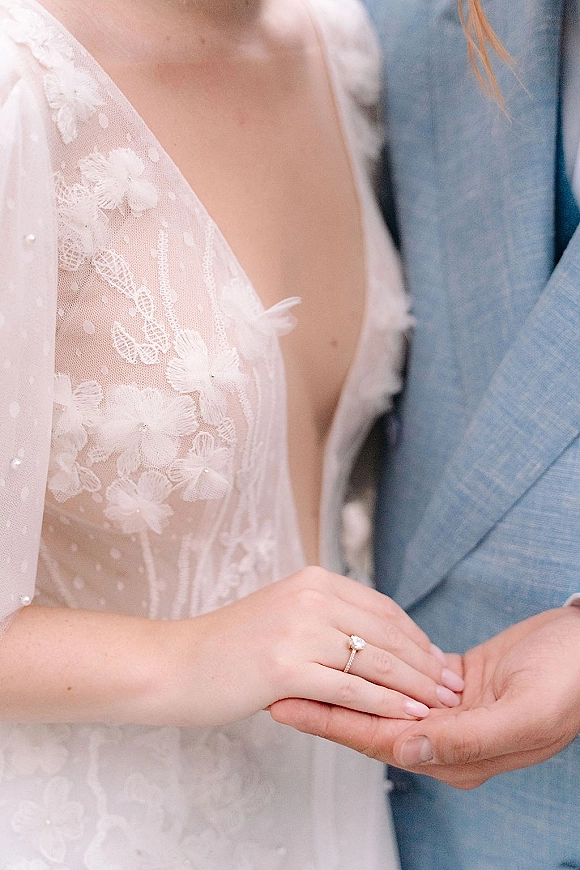 Engagement ring close-up of an oval diamond solitaire on the bride’s hand, resting on groom’s suit jacket beside lace wedding dress detail