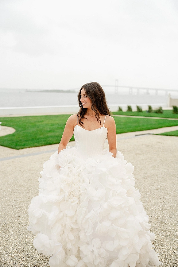 Bridal portrait of a bride in a spaghetti strap wedding dress with corset bodice and ruffled skirt by a waterfront bridge under overcast sky