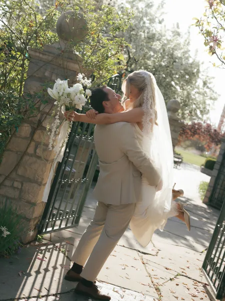 Wedding couple portrait of groom lifting bride as she holds a white bouquet with trailing ribbon by a wrought iron gate in sunlit greenery