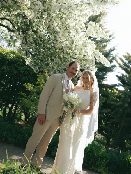Couple portrait of bride and groom posing, bride holding a white bouquet under a sunlit flowering tree on a garden path, veil flowing