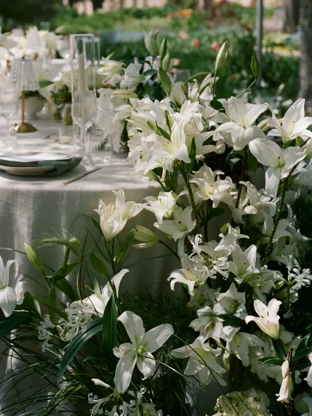 Wedding tablescape with a white lily centerpiece, greenery runner, and taper candles on white linen, set on an outdoor lawn with foliage