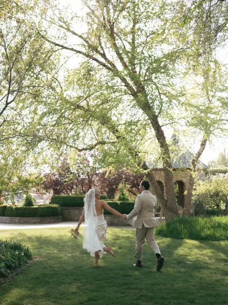 Couple portrait of bride and groom holding hands walking away on a garden lawn, bride holding heels with long veil trailing near a stone wall
