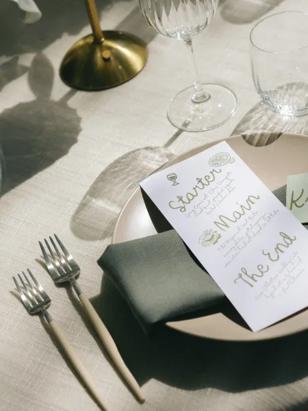 Wedding place setting with a wedding menu card on the plate, sage green napkin, place card, stemware, and brass candlestick in soft shadows