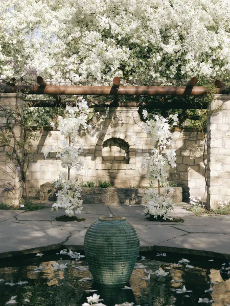 Ceremony altar with outdoor wedding altar under a wooden pergola, white floral arch and petal-filled fountain against a stone wall in a garden courtyard