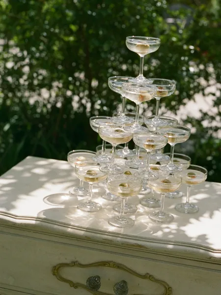 Champagne tower of coupe glasses filled with champagne and olive garnish on a white table atop a vintage dresser in sunlit garden greenery