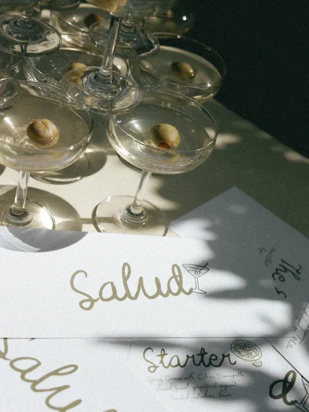 Wedding cocktail menu card with calligraphy lettering beside martini glasses and olive garnish, styled on a table with natural light shadows