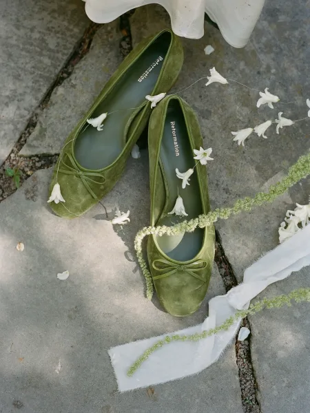 Bridal shoes in green wedding shoes velvet flats with bow, white floral sprigs and ribbon beside a wedding dress hem on stone pavement