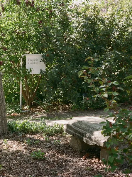 Garden seating area with a stone bench and garden sign beside a mulch path, framed by shrubs and flowers in dappled sunlight