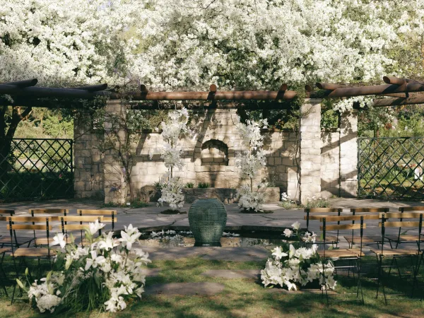 Ceremony setup for an outdoor wedding ceremony with white floral aisle clusters and chairs beneath a pergola by a stone wall fountain