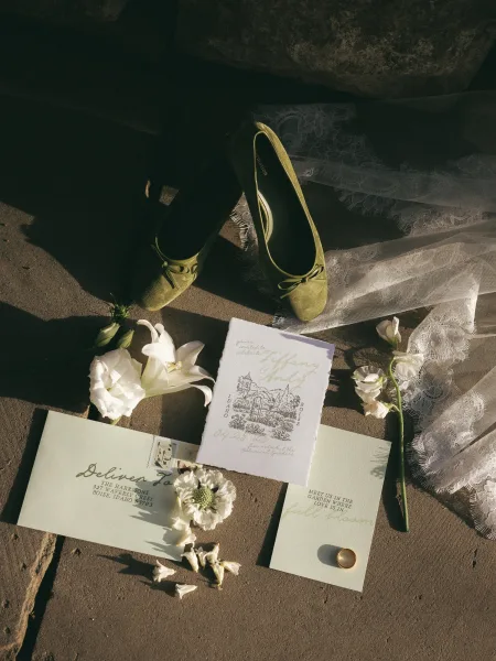 Invitation suite wedding invitation flat lay with calligraphy, wax seal, postage stamp, gold band, lace veil, green flats on stone pavement