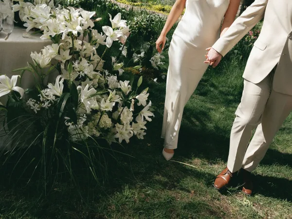 Ceremony moment as bride and groom holding hands in a garden aisle, accented by white lily florals and greenery on the grass lawn