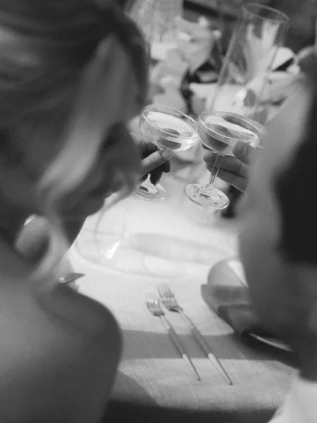 Wedding toast with champagne coupe toast as hands raise coupe glasses over a tablecloth with forks and napkin, guests blurred behind