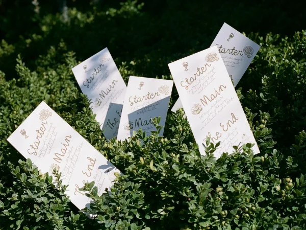 Wedding menu cards with gold ink calligraphy and small food icons, styled on a sunlit boxwood hedge with leafy shadows