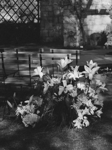 Ceremony aisle flowers with white lilies and greenery grounded on grass beside wooden folding chairs, set against a stone wall garden backdrop