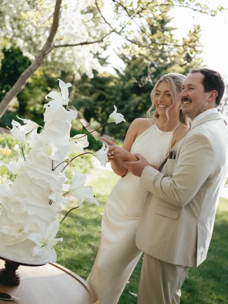 Wedding cake cutting as bride in minimalist gown and groom in beige suit hold the knife at a white cake on stand in sunny garden