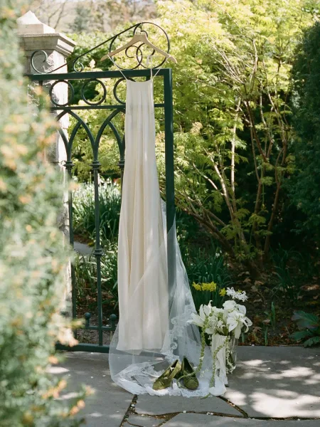 Wedding dress on hanger with lace-edged veil and lily bouquet, framed by a wrought iron gate beside garden greenery and stone path
