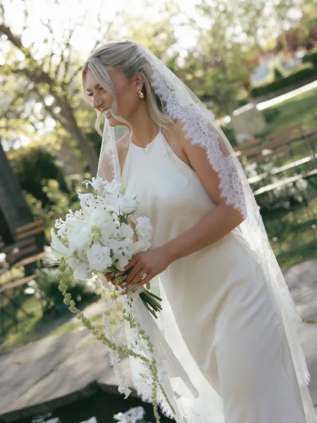 Bridal portrait of a smiling bride holding a white bouquet, lace veil draped over her shoulders in a garden with trees and stone walkway