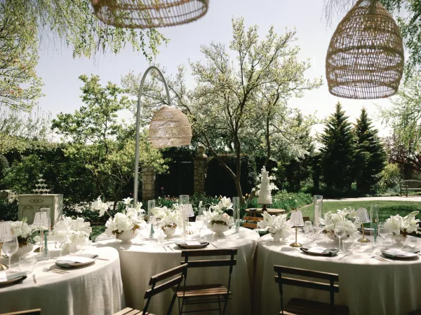 Reception tablescape with round linen tables, white floral centerpieces, taper candles and small lamps under wicker pendant lights on a garden lawn