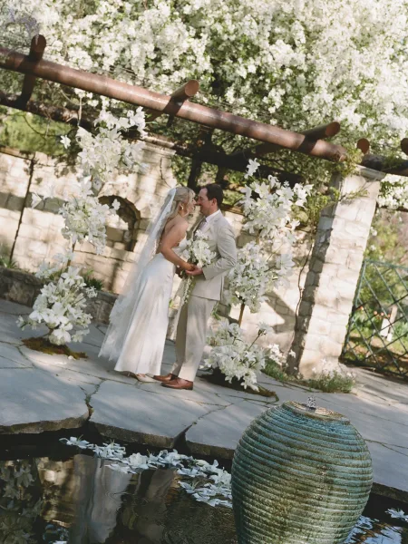 Wedding couple portrait of bride and groom by pond, touching foreheads under a pergola, bride holding bouquet with long veil and stone wall behind